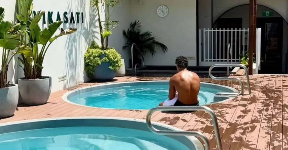 A man sits by a circular pool at a Gold Coast bathhouse, near potted plants and a wall clock.