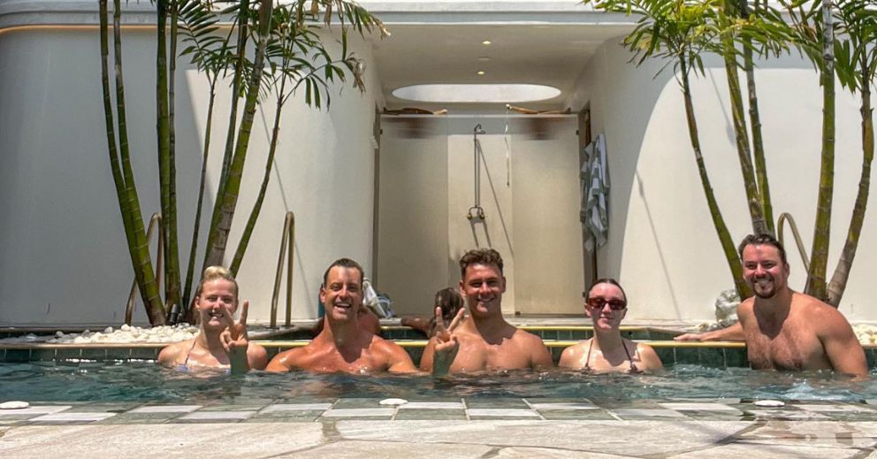 Five people smile in a pool during their first bathhouse visit in Brisbane with palm trees behind.