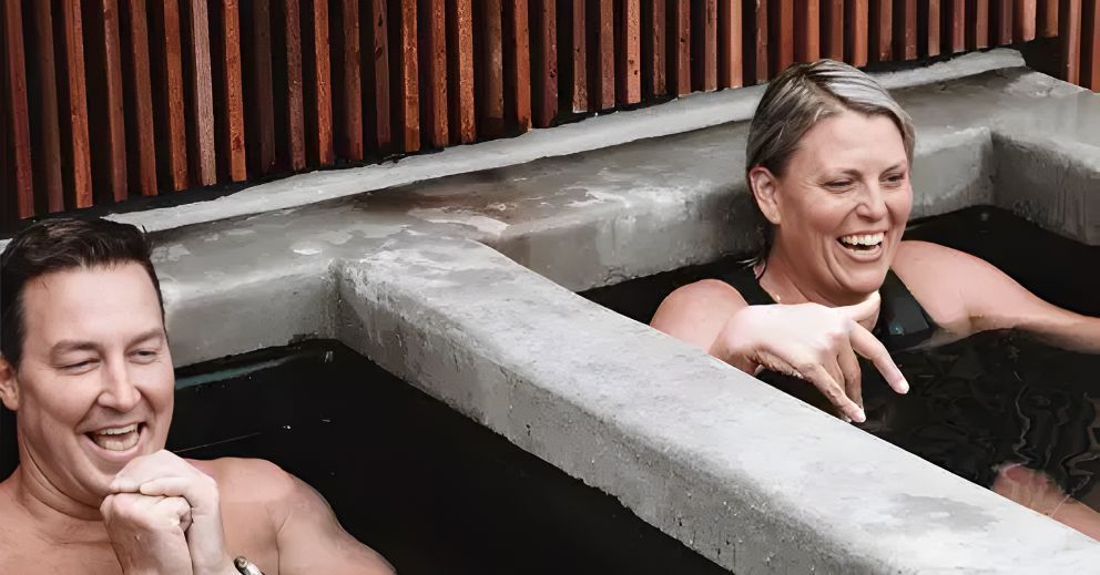 Two adults smiling and talking at a Brisbane bathhouse in adjacent concrete hot tubs.