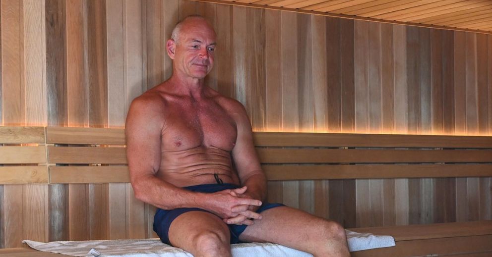 A shirtless man in swim trunks relaxes on a towel inside a sauna, boosting heart health.