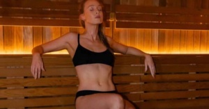 Woman in a black bikini sits relaxed on a wooden bench, embracing sauna hydration and warm lighting.