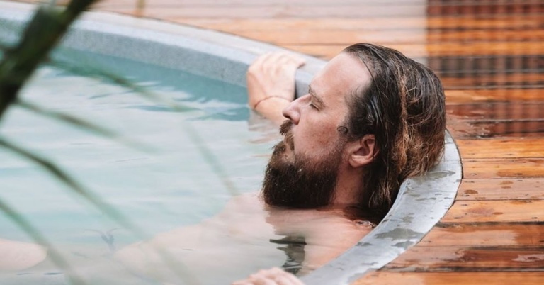 Man with long hair and beard relaxing in a round outdoor pool—learn how to avoid dryness after swimming.