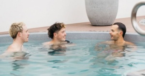 Three men chat in a round magnesium pool, easing posture pain; a planter sits in the background.