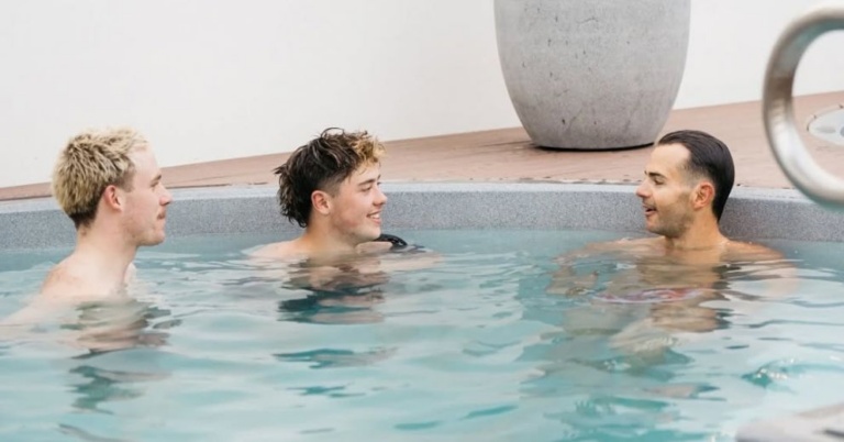 Three men chat in a round magnesium pool, easing posture pain; a planter sits in the background.