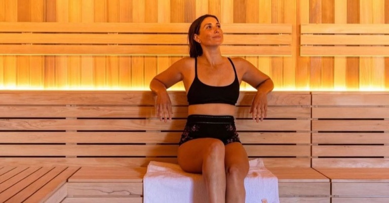 Woman practicing heat adaptation, sits on a towel in a wooden sauna, wearing black sportswear.