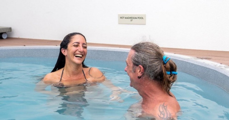 Two people smiling and talking while relaxing in a Magnesium Pool Recovery outdoor pool with a sign.
