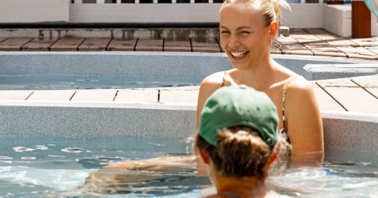 A smiling woman in a hot tub next to a person in a green baseball cap, knowing what to bring.